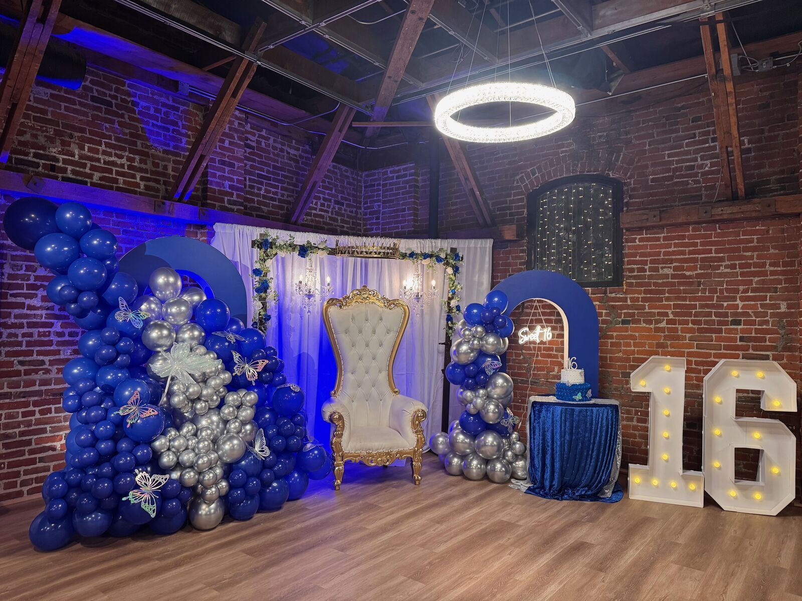 Sweet 16 setup at The Prestige Hall with blue and silver balloon arch, throne chair, and marquee lights against exposed brick walls